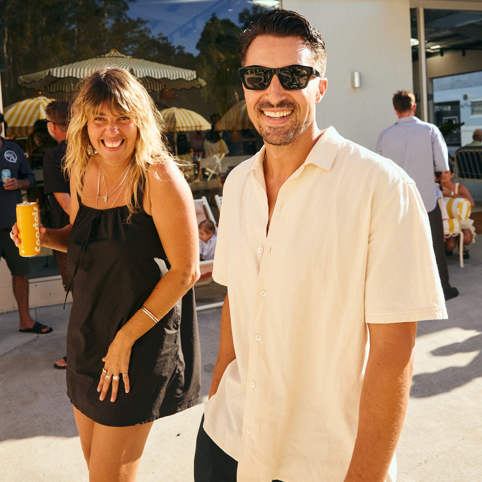 Man and woman standing together outdoors, smiling, holding a Pineapple Coastals can, with a building and people in the background.