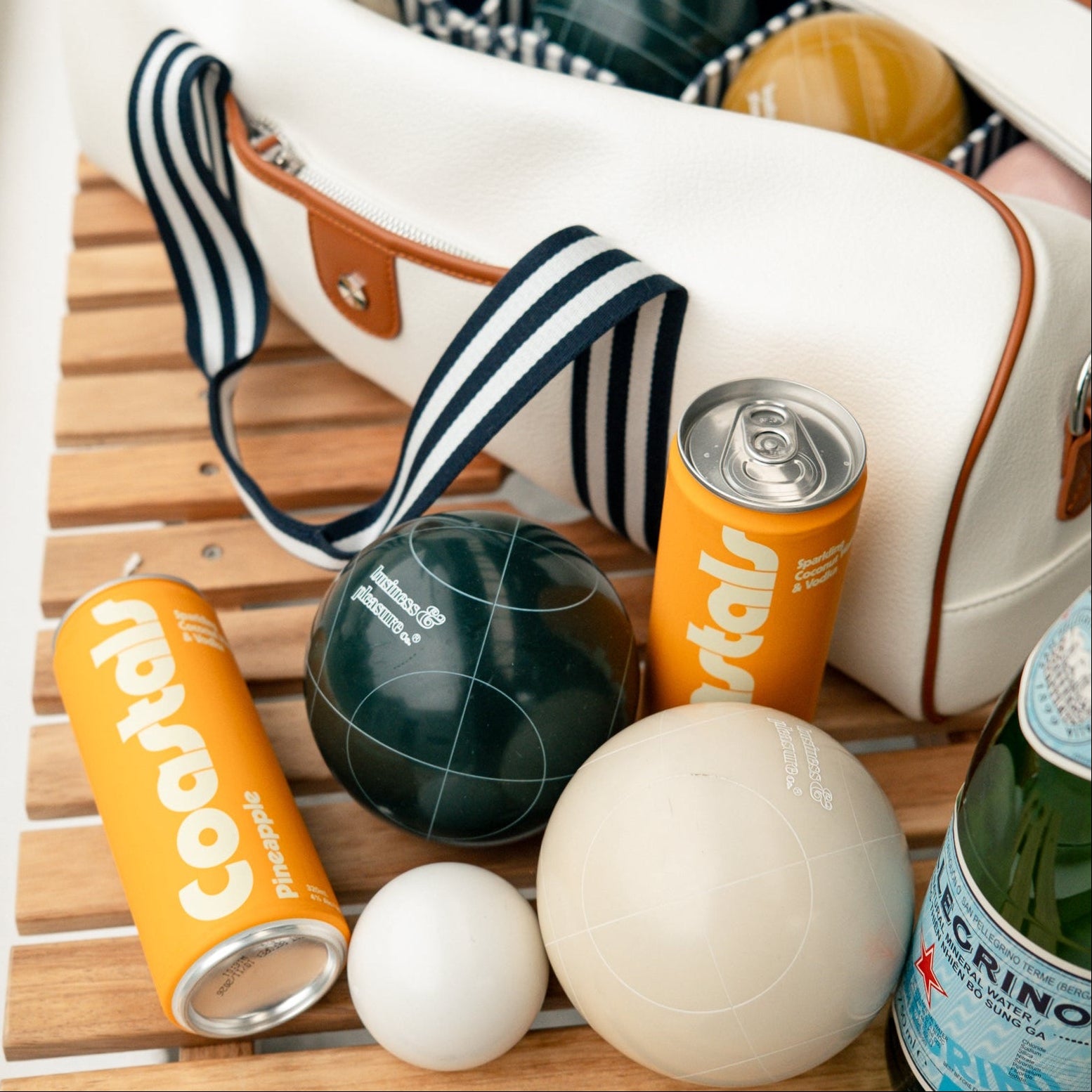 Close-up of a white bowls bag with items including a Coastals Pineapple can and bowls balls on a wooden surface.