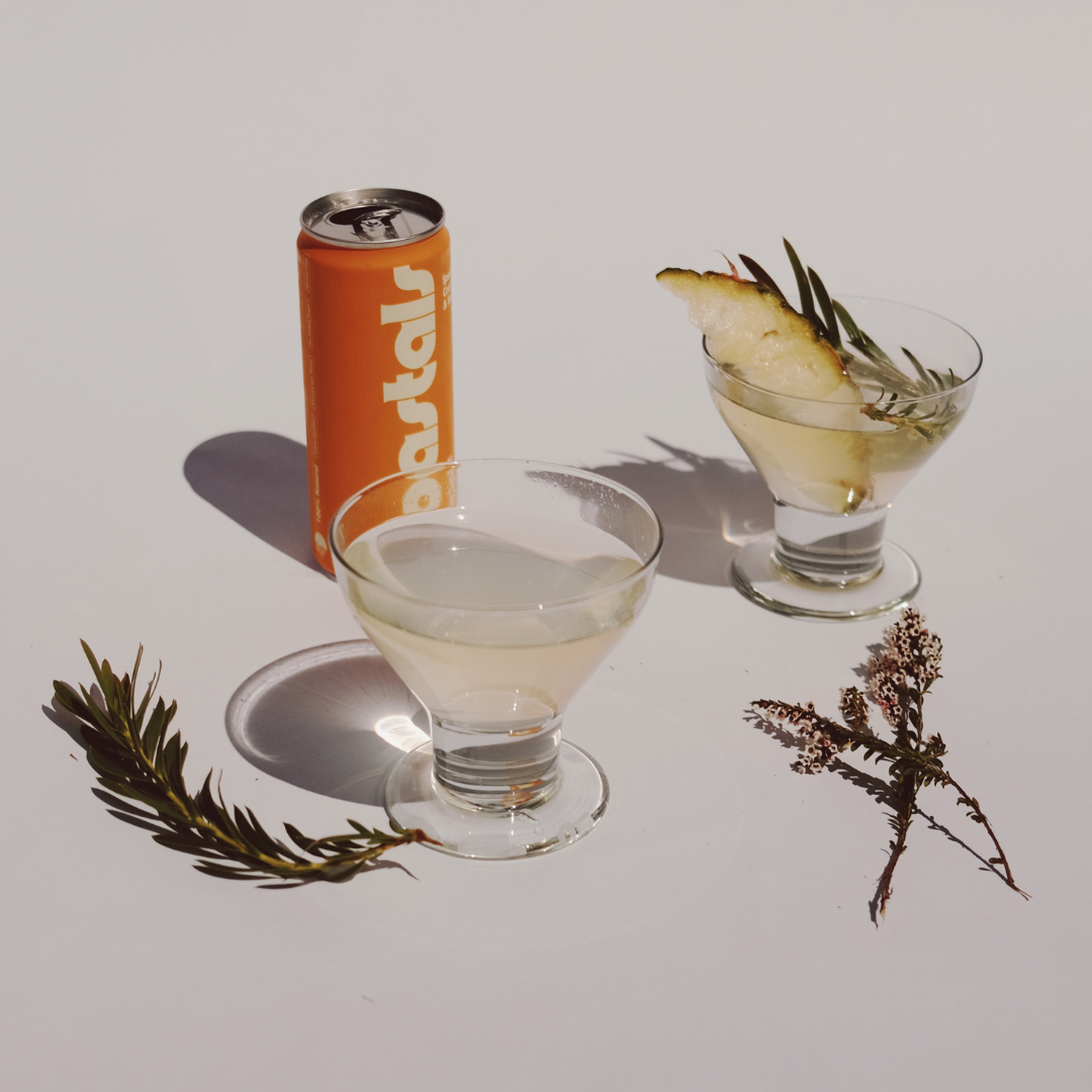 Coastals Pineapple can beside two cocktails garnished with pineapple and herbs on a white background.
