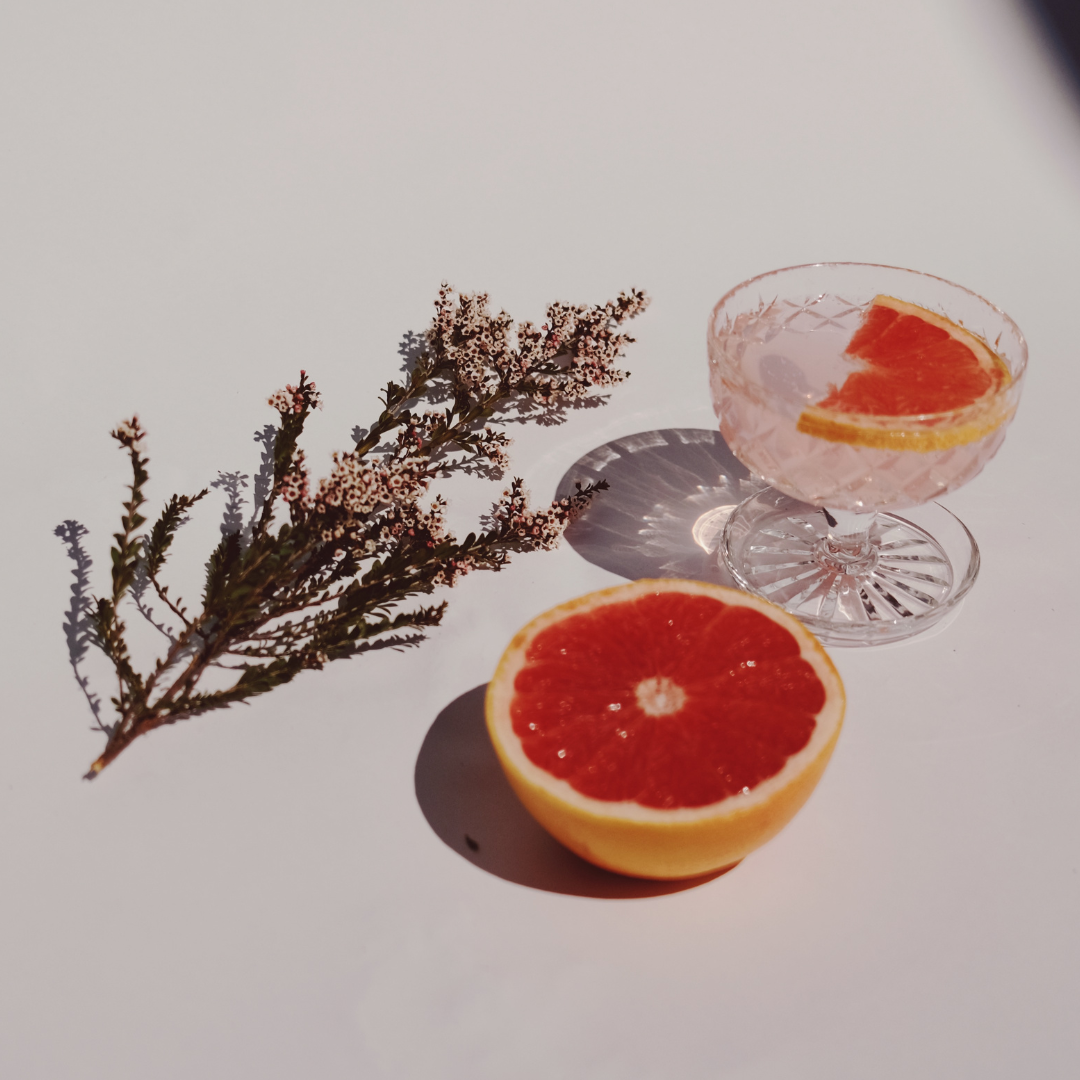Pink grapefruit cocktail in a cut-glass coupe with a grapefruit slice and sprig of tiny white flowers on a white background.